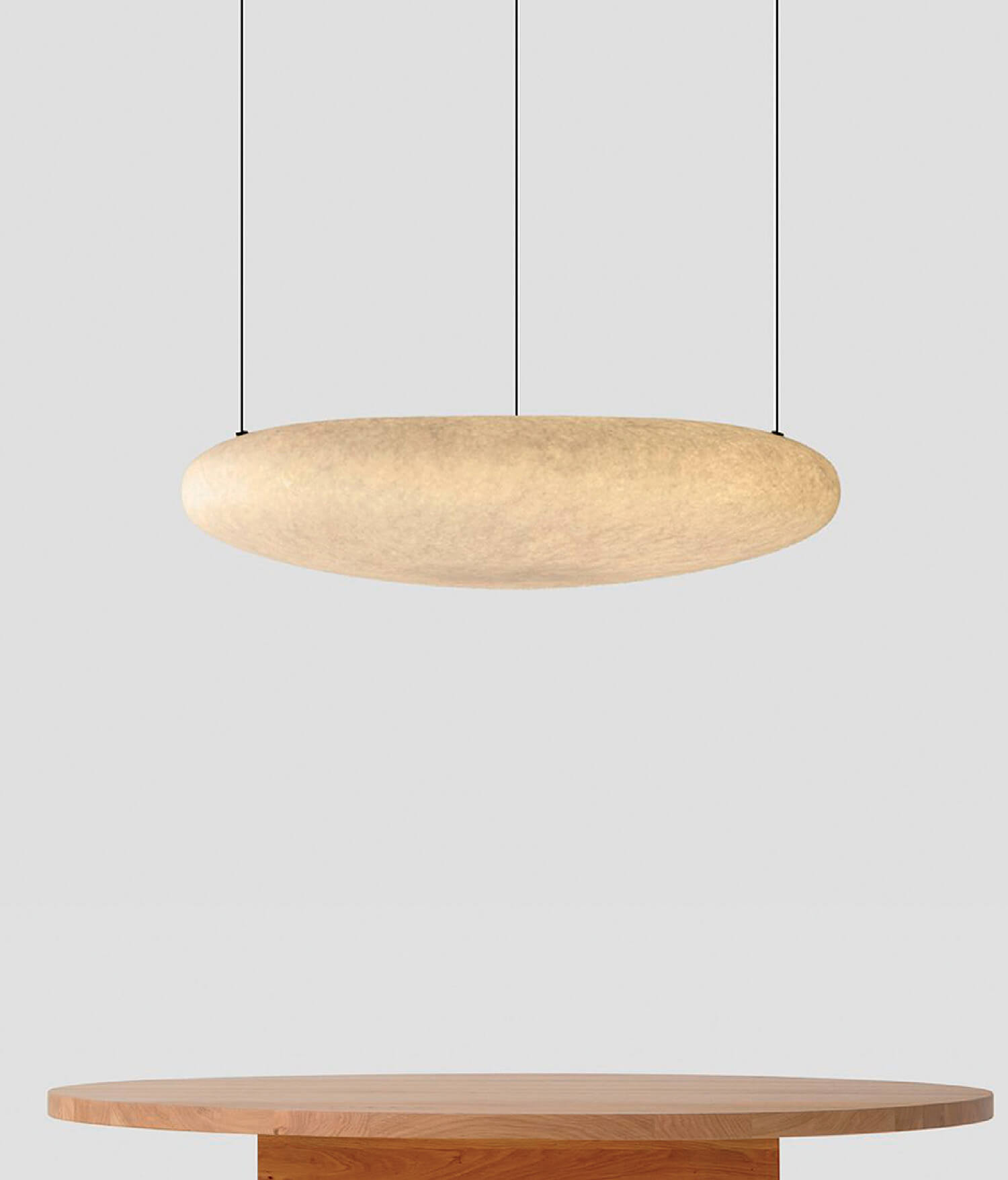 A round wooden table beneath a large, disk-shaped pendant light suspended by three cables against a plain gray background.