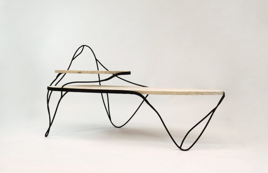A bench with a light wooden seat and shelf, supported by thin, wavy black metal legs and frame, set against a plain white background.