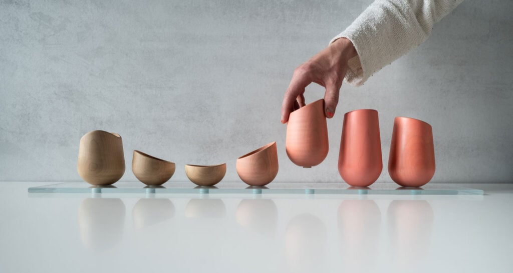 A hand places a reddish-brown ceramic cup among a row of wooden and ceramic cups on a glass shelf against a gray background, capturing the artisanal spirit of London Craft Week.