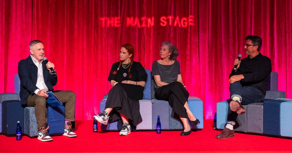 Four people sit on stage in front of a red curtain labeled "The Main Stage," engaged in a panel discussion with microphones and water bottles.