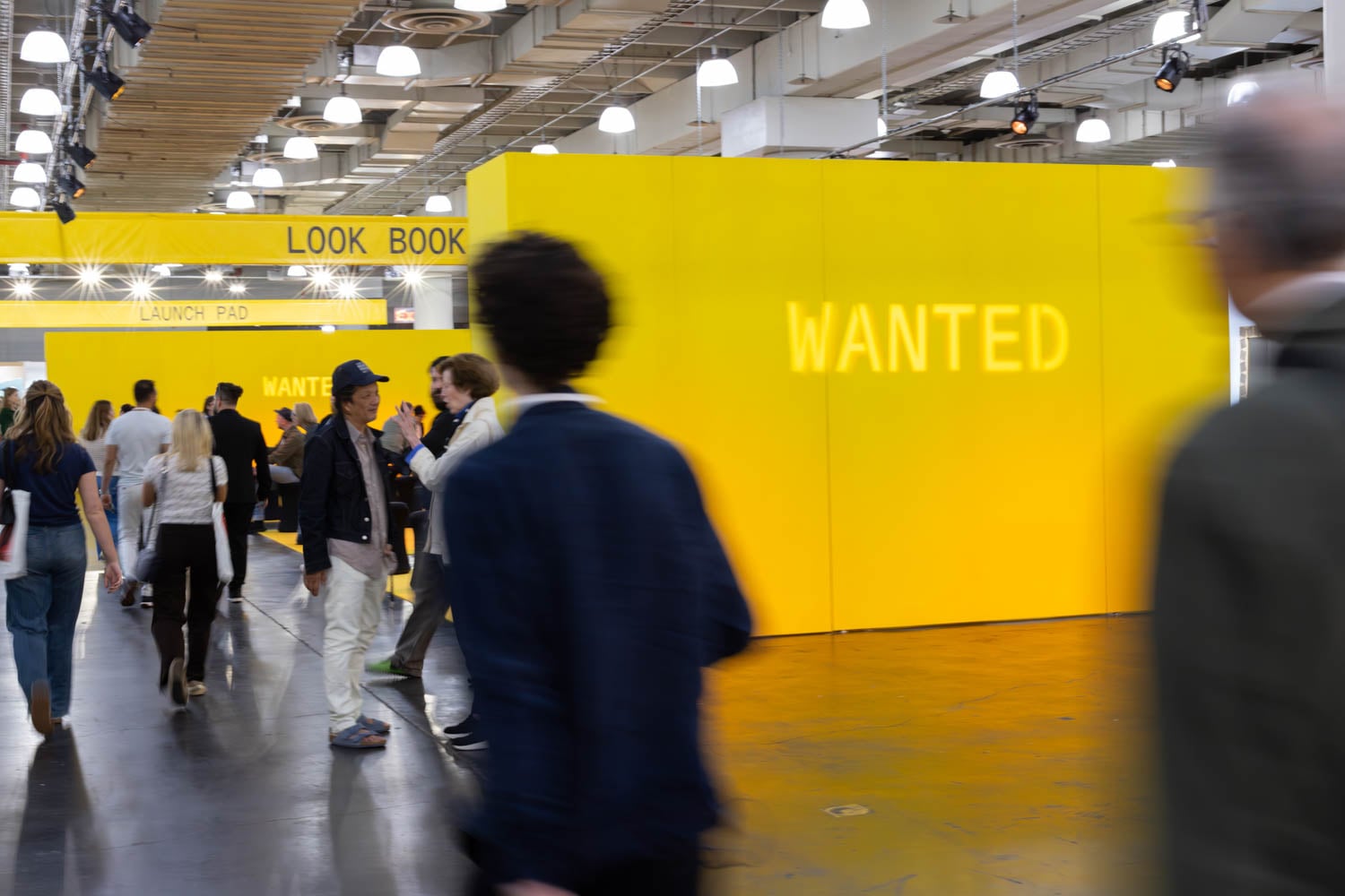 People walk through an indoor event space with bright yellow walls displaying the words "LOOK BOOK," "LAUNCH PAD," and "WANTED" in large letters.