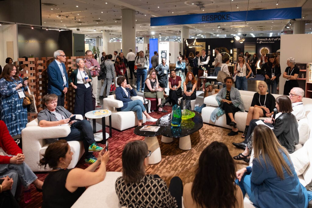 A group of people sit in a circle on white sofas and chairs at an indoor event space, while others stand and observe; exhibition booths are visible in the background.