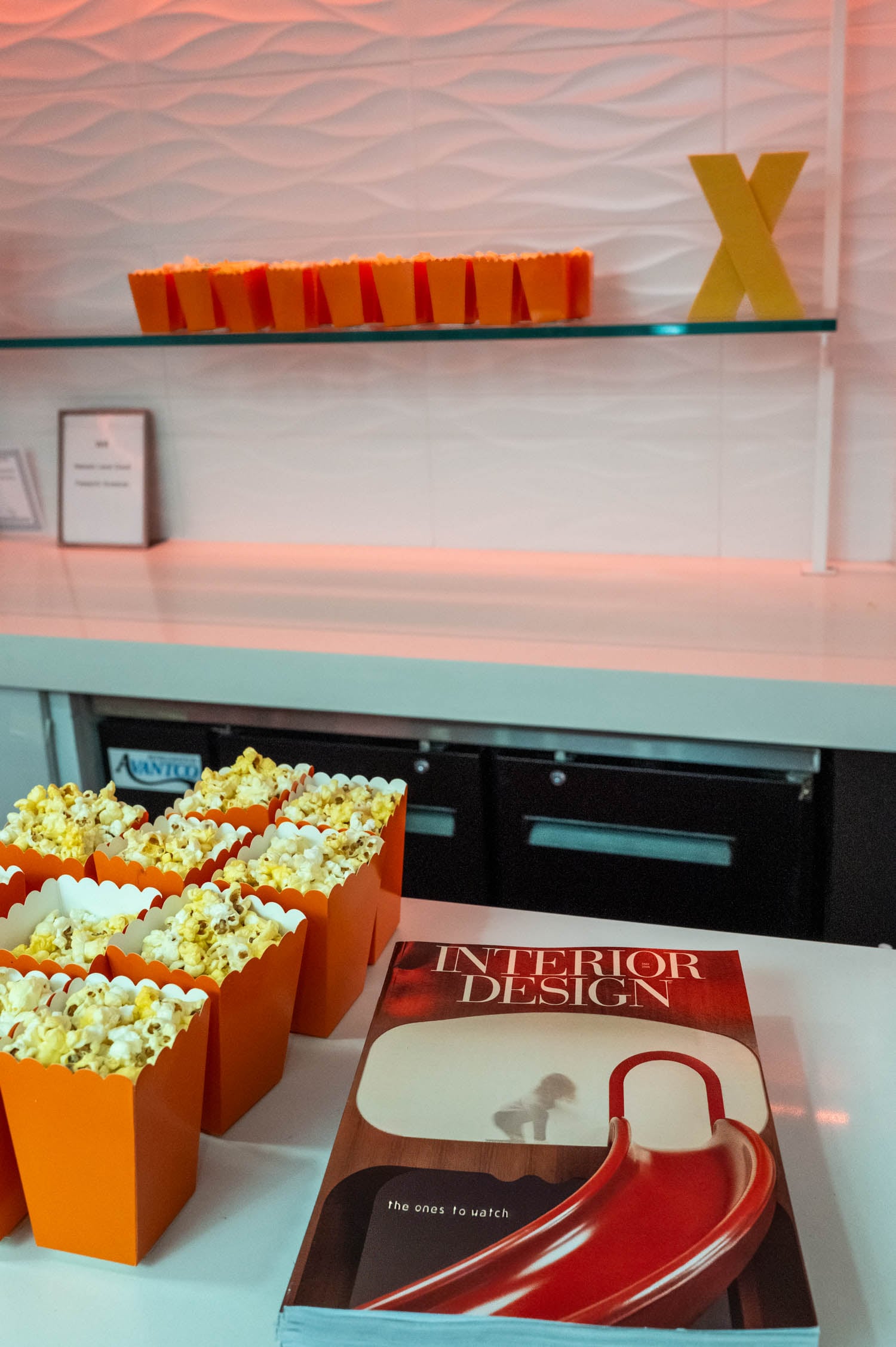 Several orange popcorn containers sit on a counter next to a magazine titled "Interior Design," celebrating the NYCxDESIGN Awards, with more popcorn containers and a yellow "X" on a shelf in the background.