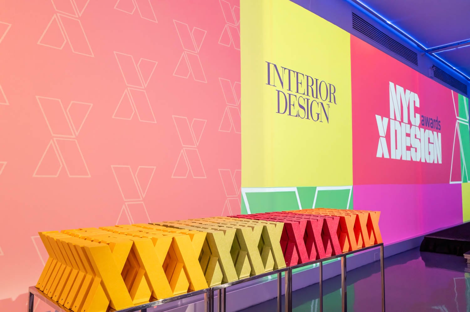 Colorful X-shaped awards are lined up on a table in front of a geometric backdrop, proudly displaying the “Interior Design” and “nycxdesign awards” logos.