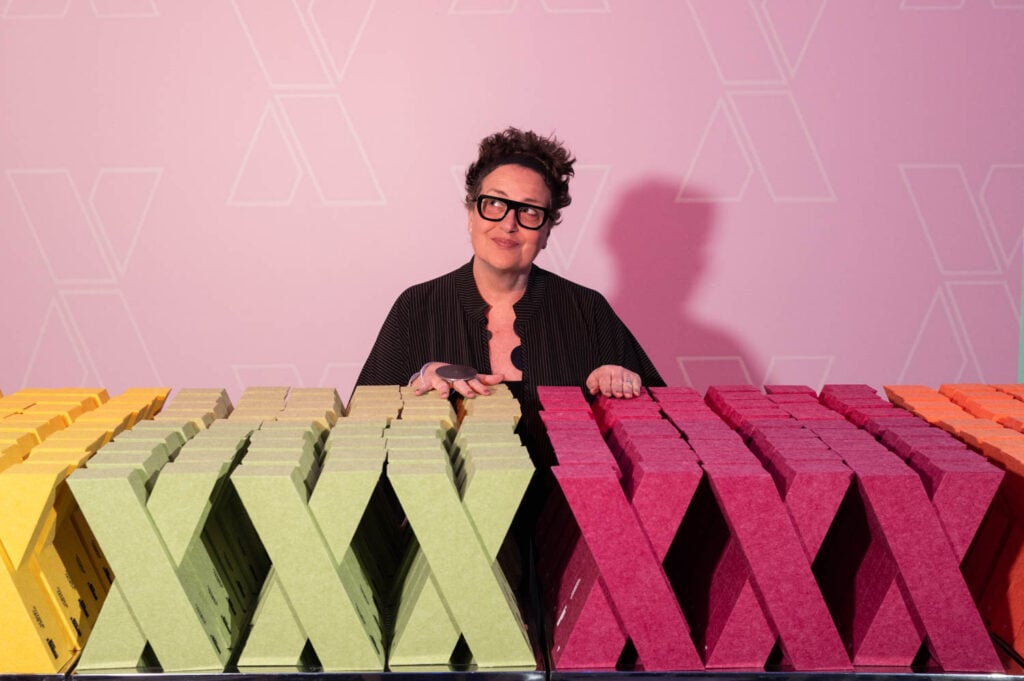 A person with short hair and glasses stands behind rows of yellow, green, and pink X-shaped objects against a pink background with X patterns, evoking the creative spirit of the NYCxDESIGN Awards.