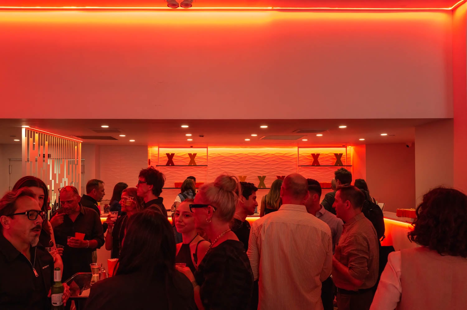 A group of people socialize in a modern, NYCxDESIGN Awards-worthy bar with red ambient lighting and decorative plants on shelves behind the bar counter.