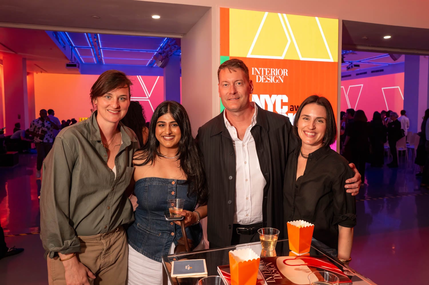 Four people stand together at an event, posing and smiling for the camera, with drinks and snacks on the table in front of them at the brightly lit NYCxDESIGN Awards gathering.