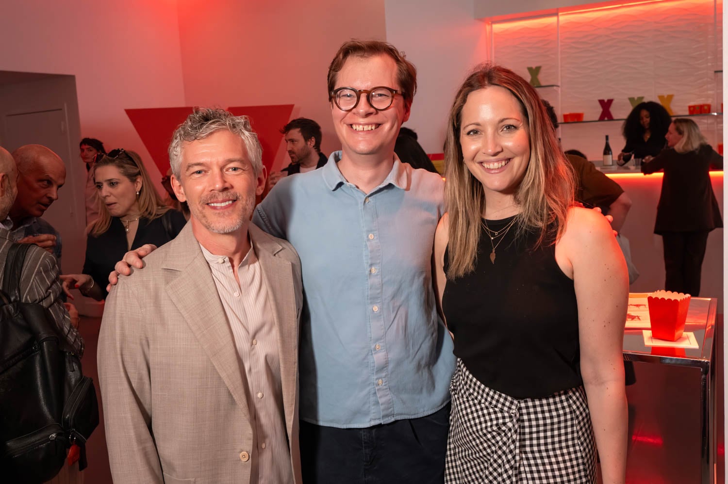 Three people stand close together and smile at the camera in a brightly lit room with a red and white color scheme during the NYCxDESIGN Awards, while others socialize in the background.