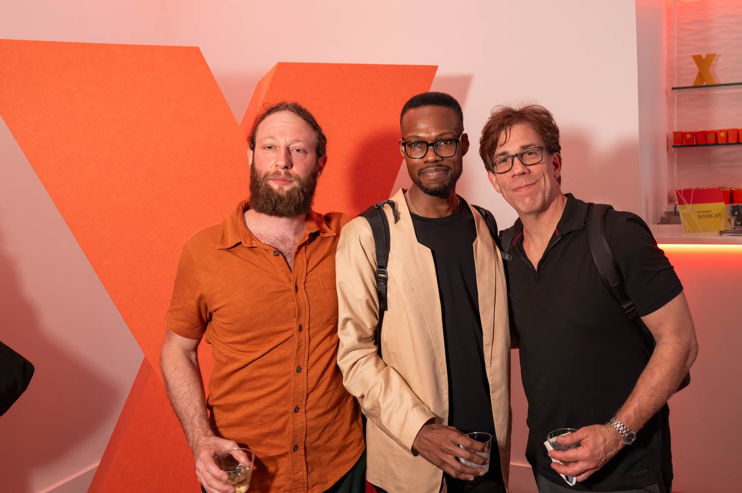 Three men stand together holding drinks, posing for a photo at an event with a large orange "X" and modern decor in the background, celebrating at the NYCxDESIGN Awards.