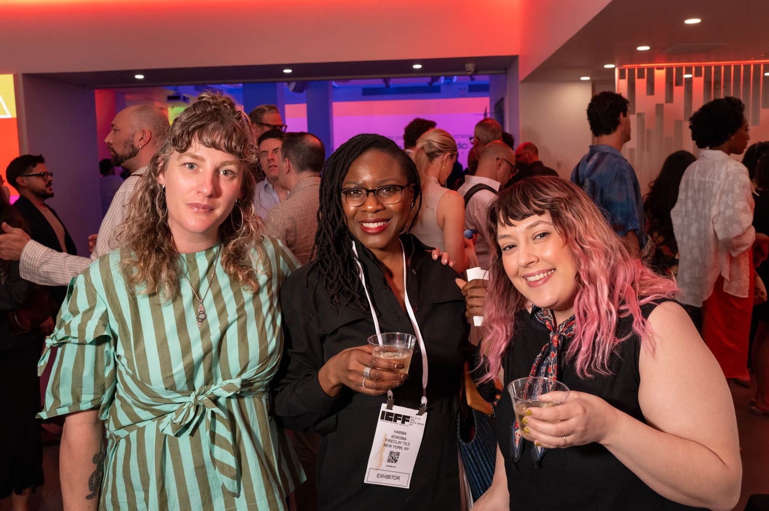 Three people stand together holding drinks at a lively indoor event, possibly celebrating the NYCxDESIGN Awards, as other attendees mingle in the background under colorful lighting.