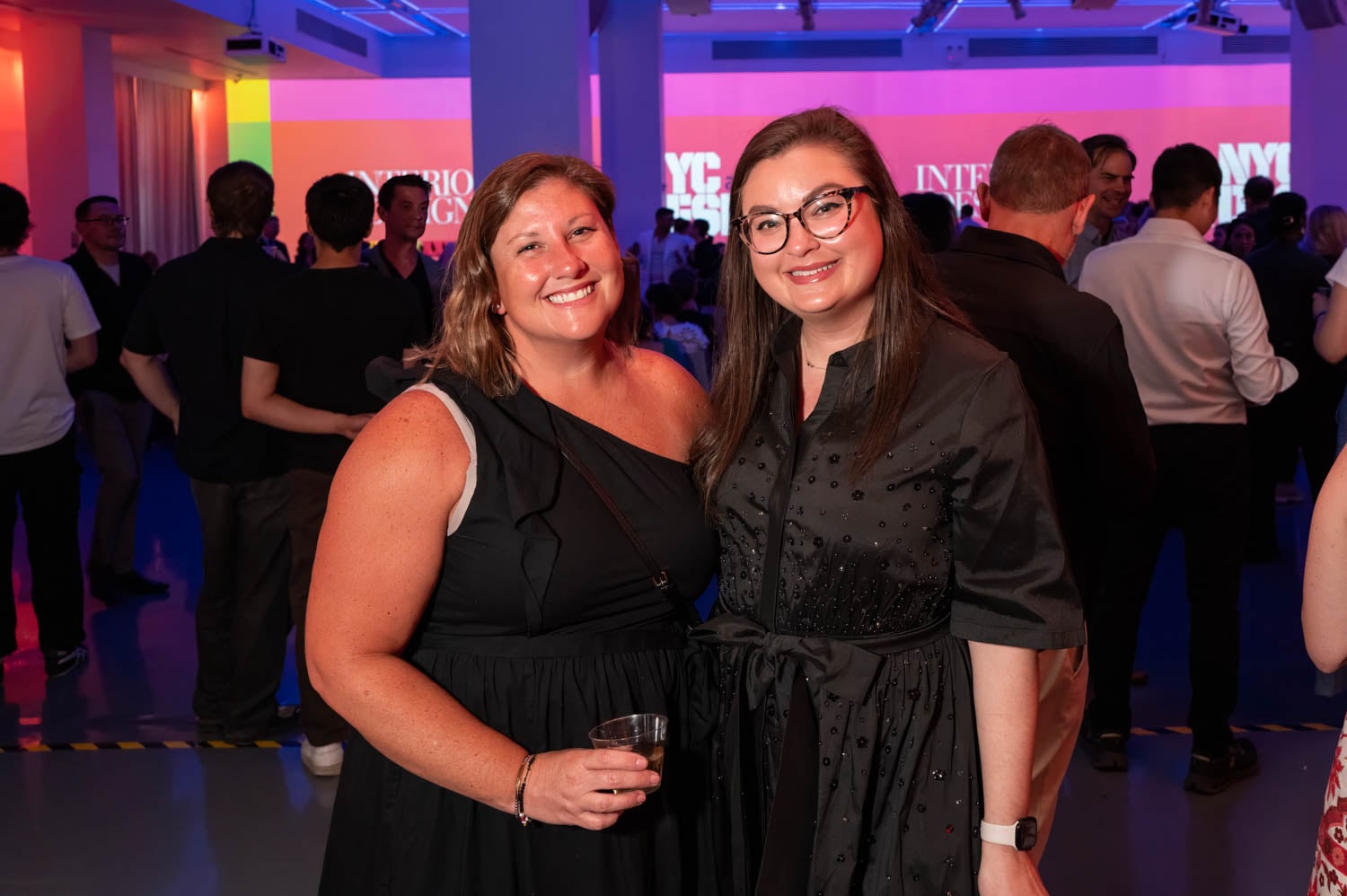 Two women pose and smile at an indoor event with colorful lighting during the NYCxDesign Awards. Other attendees are visible socializing in the background.