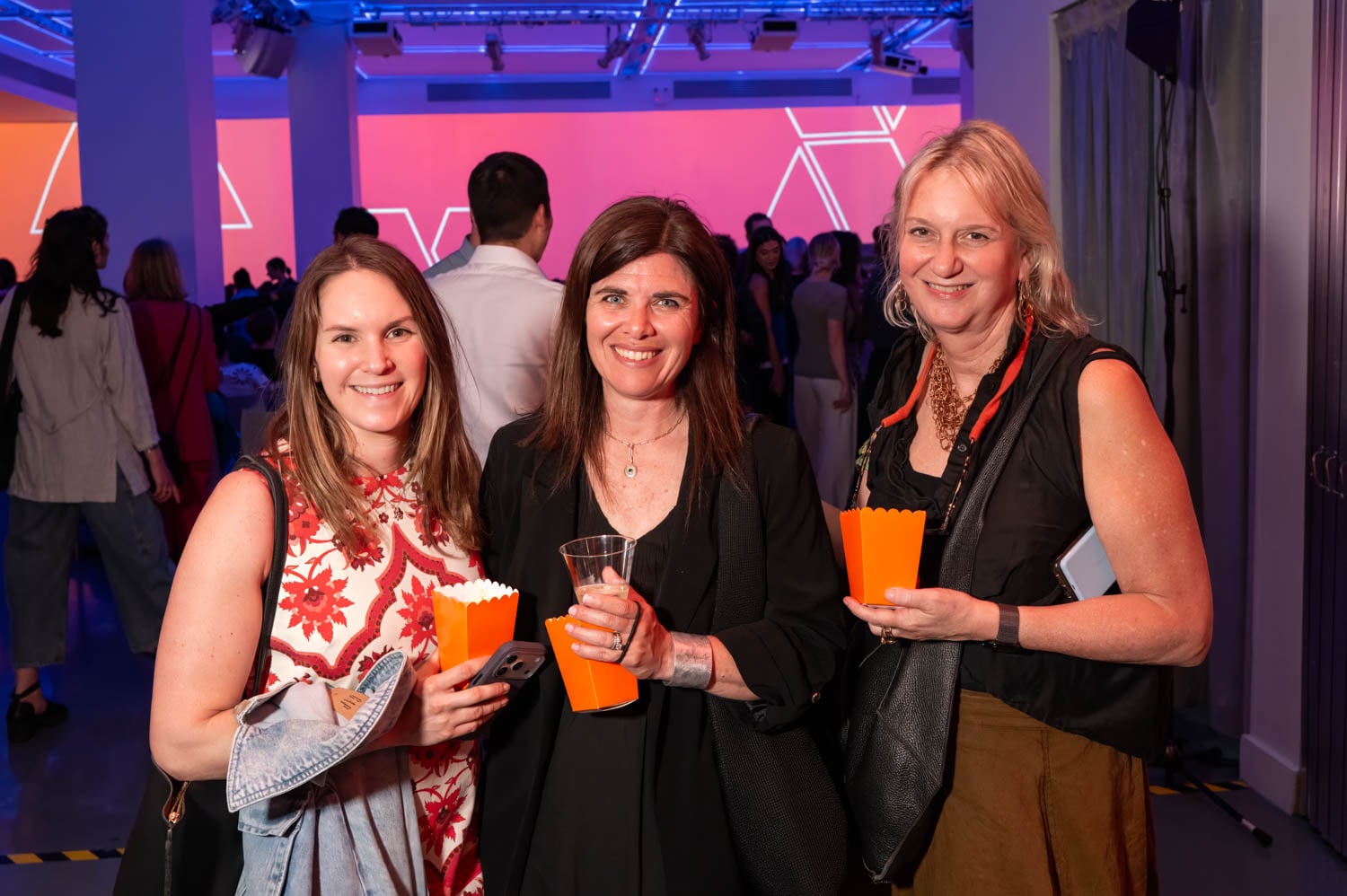 Three women standing together indoors at an event, holding orange popcorn containers and smiling at the camera during the NYCxDESIGN Awards, with other attendees and colorful lighting in the background.