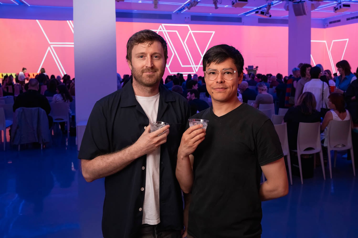 Two people stand together holding drinks at an indoor event, with a crowd seated in the background and pink lighting on the walls, capturing the vibrant atmosphere of the NYCxDESIGN Awards.