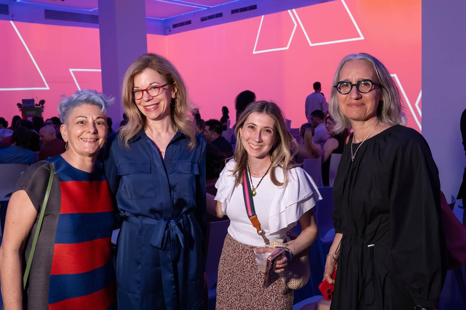 Four women stand together and smile for a photo at an indoor event, with vibrant pink and purple geometric lighting in the background at the NYCxDesign Awards.