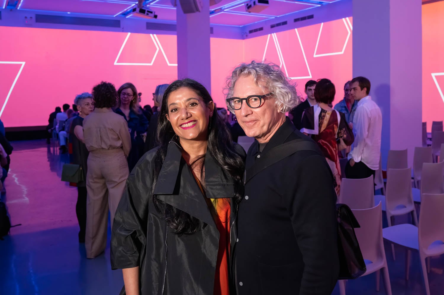 Two people pose together and smile for a photo at an indoor event, likely the NYCxDesign Awards, with other attendees and geometric pink lighting in the background.