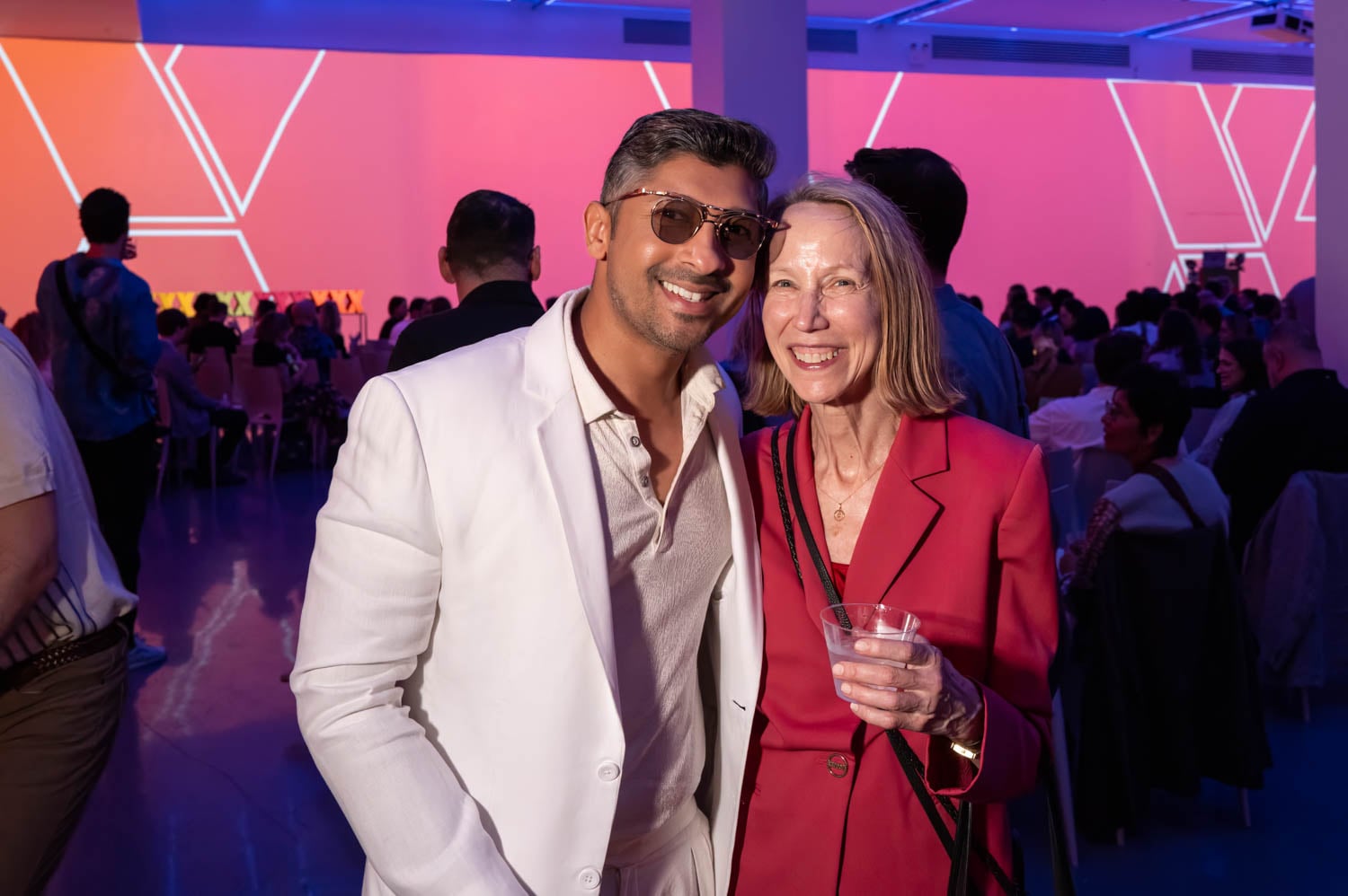 Two people smiling and posing together at an indoor event with a colorful, geometric-lit background and a seated audience behind them, celebrating the excitement of the NYCxDESIGN Awards.