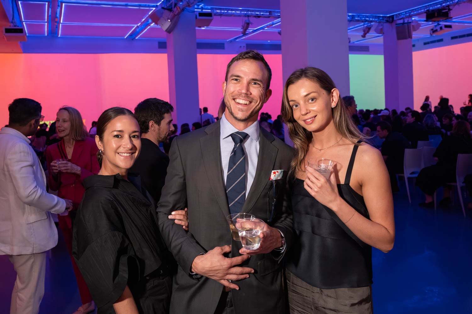 Three people stand close together and smile at the camera, holding drinks at the nycxdesign awards, a formal indoor event with colorful lighting and a seated audience in the background.