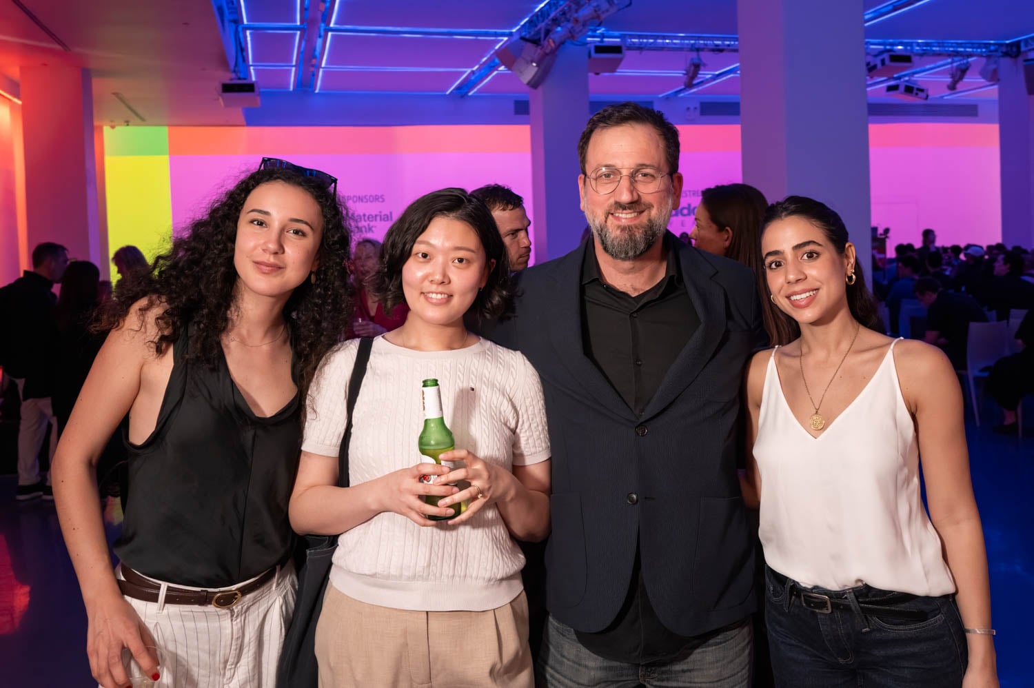 Four people posing together at an indoor event with colorful lights and a crowd in the background; one person is holding a drink at the NYCxDesign Awards.