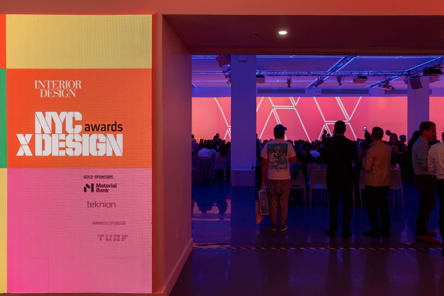 A group of people attends the NYCxDESIGN Awards event in a large, brightly lit room filled with geometric light displays and an eye-catching nycxdesign awards sponsor sign at the entrance.