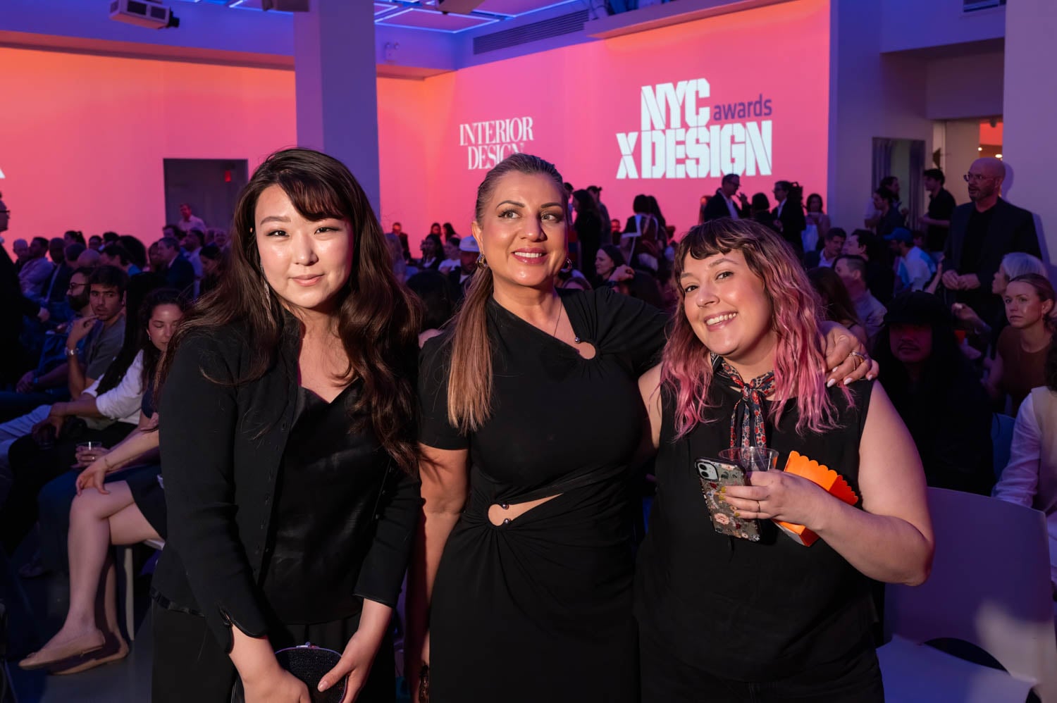 Three women pose for a photo at an indoor event, with seated attendees and "NYCxDESIGN Awards" projected on the wall in the background.