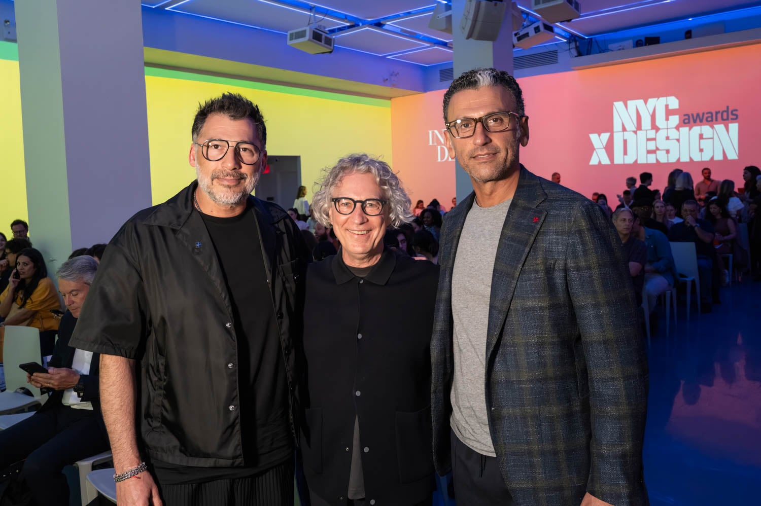 Three men stand together and pose for a photo at an indoor event, with the NYCxDESIGN Awards logo projected in the background and audience members seated nearby.