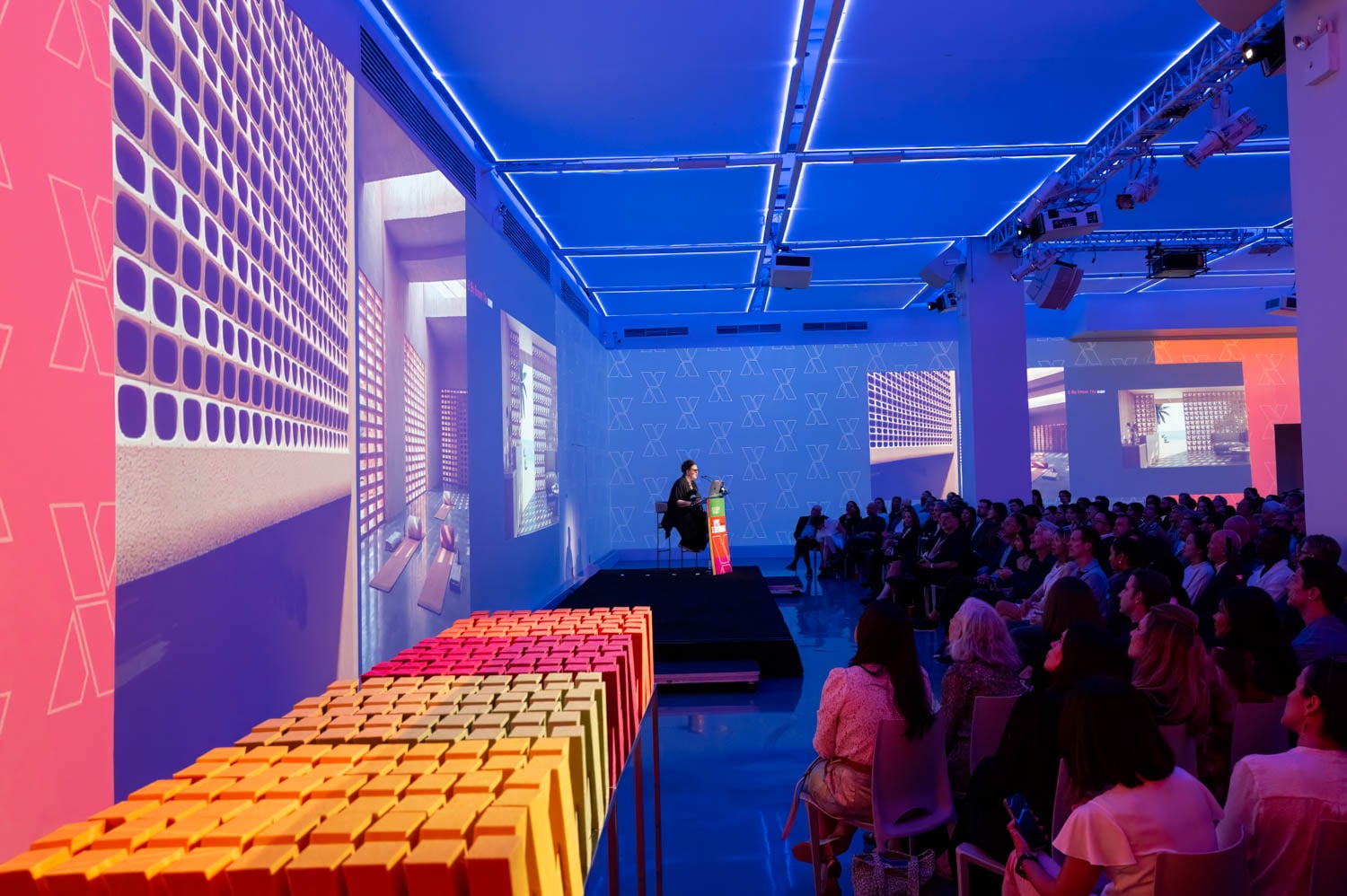 A speaker presents on stage at the NYCxDesign Awards in a modern, blue-lit event space with large screen projections and rows of colorful boxes in the foreground.