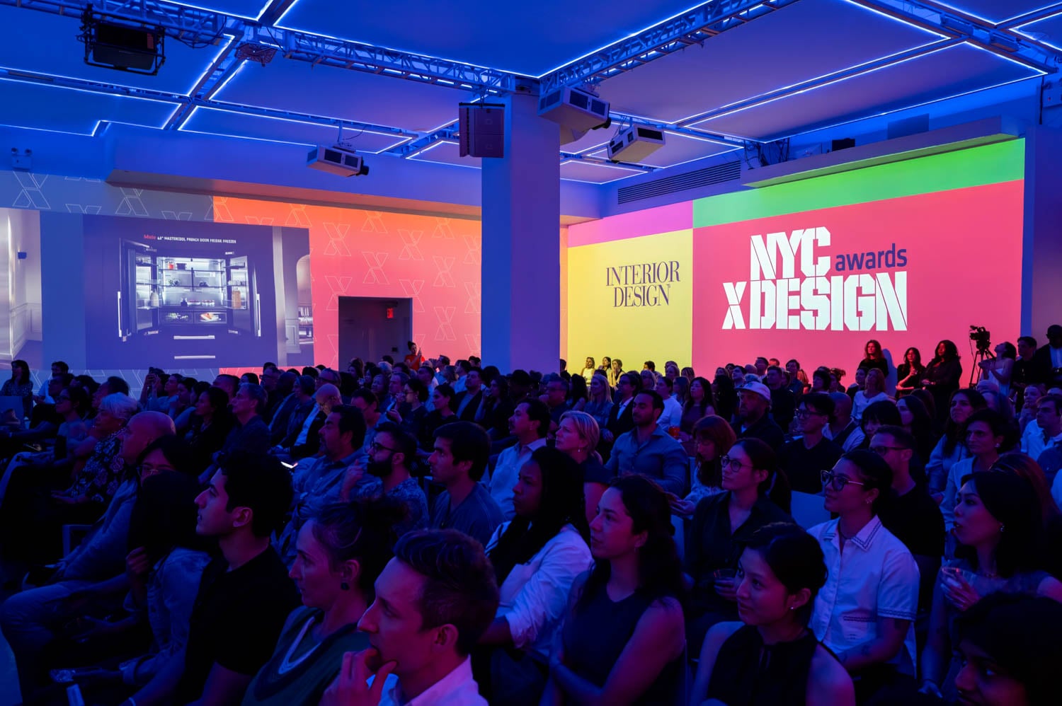 A seated audience watches a presentation at the NYCxDESIGN Awards event, surrounded by colorful lighting and large projected NYCxDESIGN Awards logos illuminating the walls.