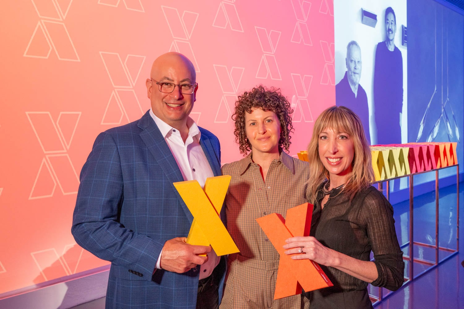 Three people stand indoors holding large, colorful X shapes, with a pink and orange background featuring repeated X patterns and a projected image—celebrating the creative spirit of the NYCxDESIGN Awards.
