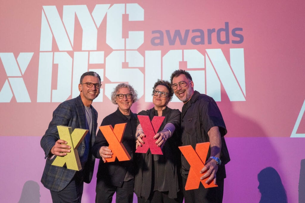 Four people stand in front of an NYCxDESIGN awards backdrop, each proudly holding a large "X" trophy and smiling at the camera, celebrating their NYCxDESIGN awards achievement.