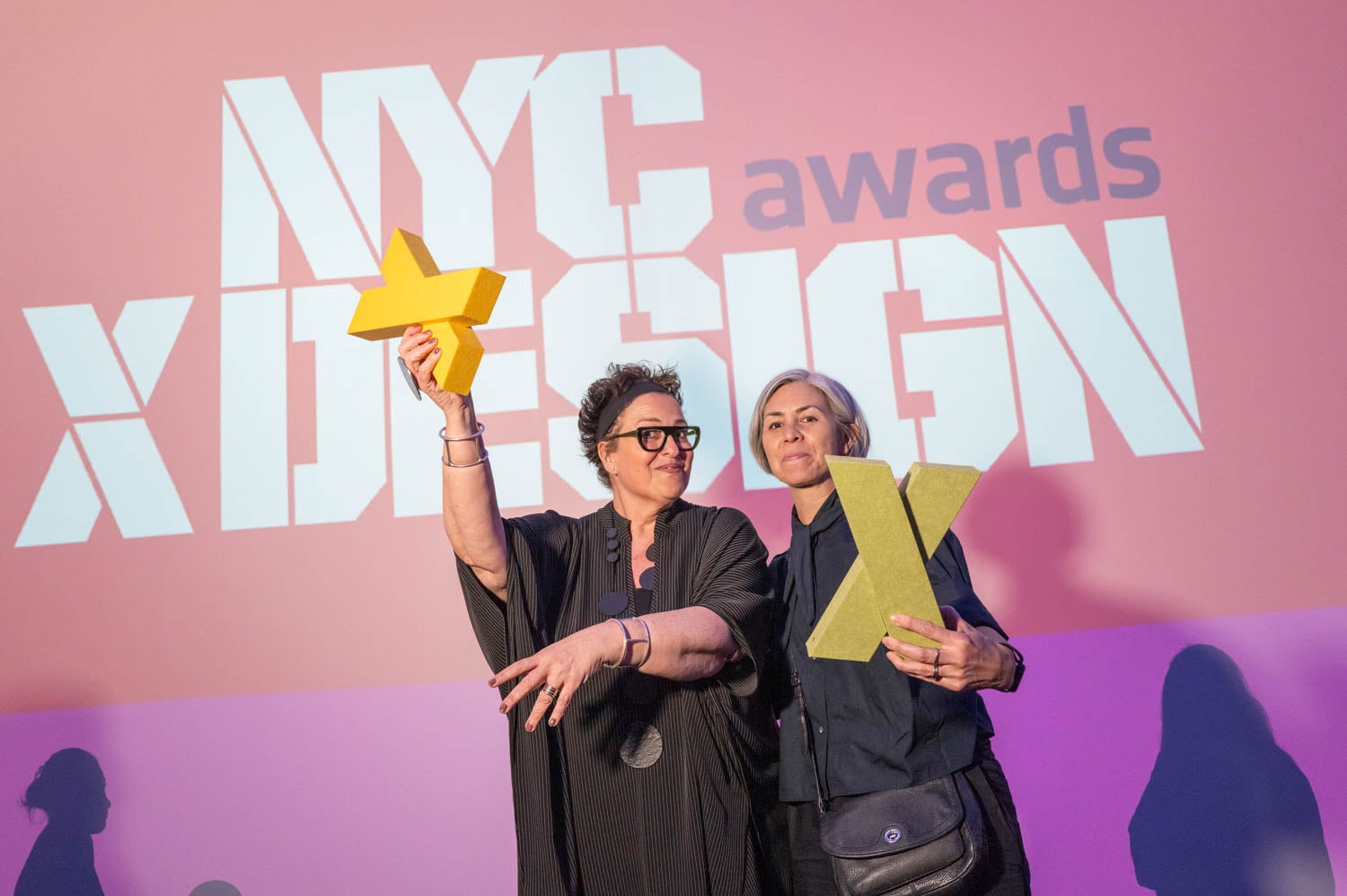 Two women hold yellow awards in front of a large NYCxDESIGN Awards sign projected on a pink and purple wall, celebrating their achievements at the NYCxDESIGN Awards event.