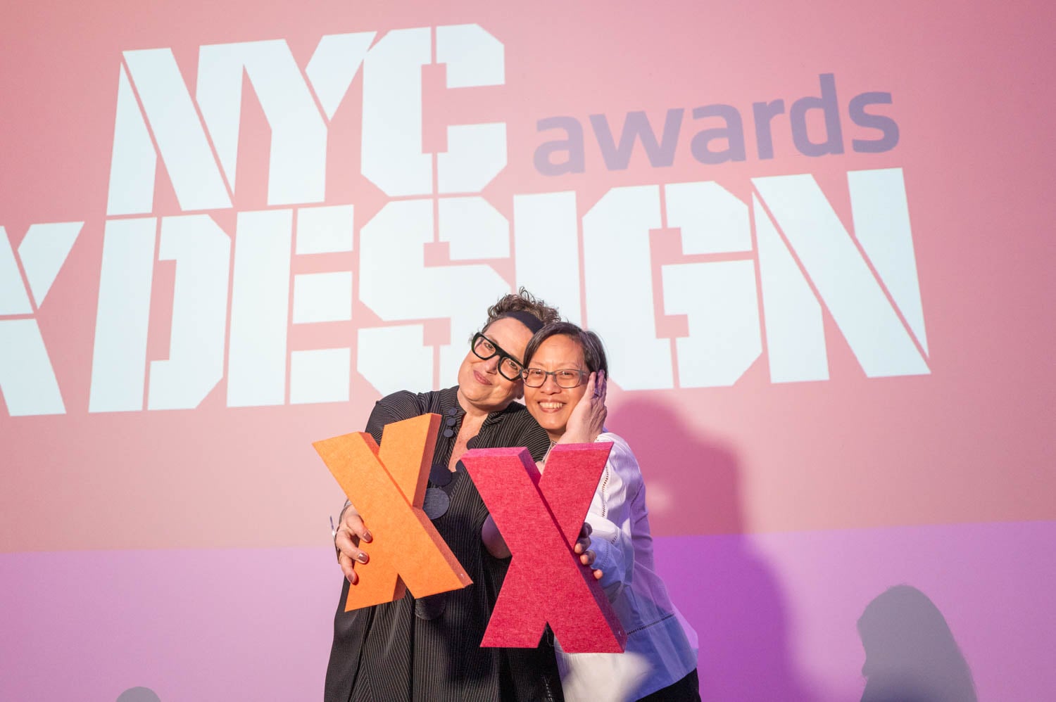 Two people pose together holding large red "X" props in front of a screen displaying the nycxdesign awards.