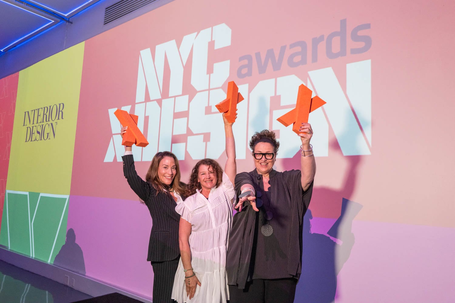 Three women stand in front of a NYCxDESIGN Awards backdrop, smiling and raising their orange NYCxDESIGN Awards in celebration.