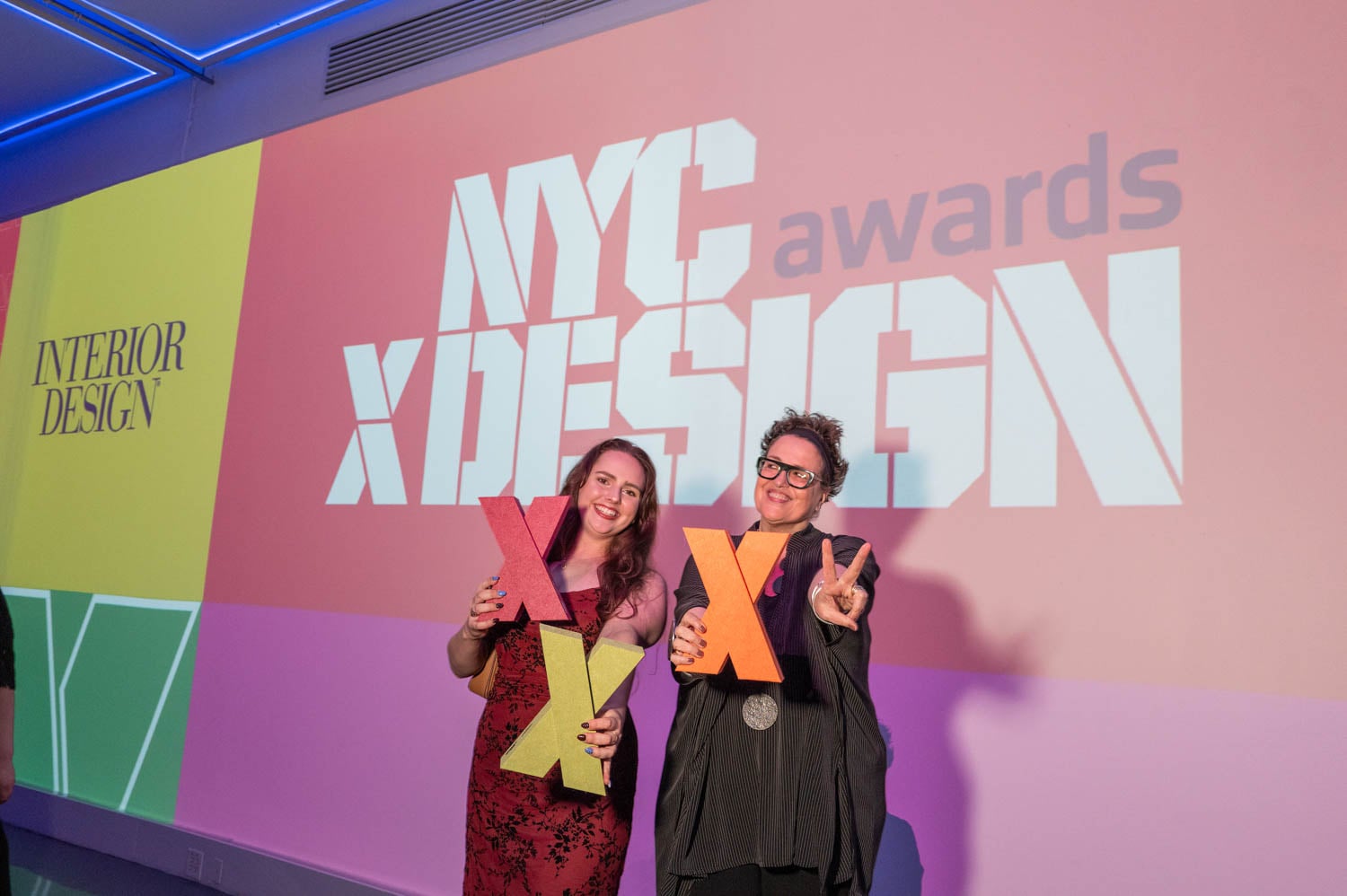 Two people stand in front of a large NYCXDESIGN awards display, holding colorful X-shaped props and smiling at the event.
