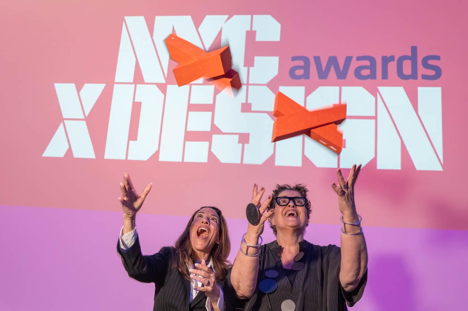 Two people in business attire throw red airplane-shaped objects in front of a screen displaying "NYCxDESIGN Awards," celebrating the spirit of innovation at the NYCxDESIGN Awards event.