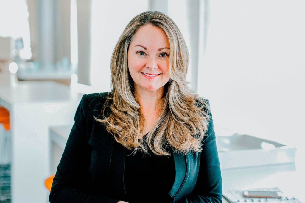 A woman with long, wavy blonde hair wearing a black blazer is smiling while seated in a bright, modern office setting.
