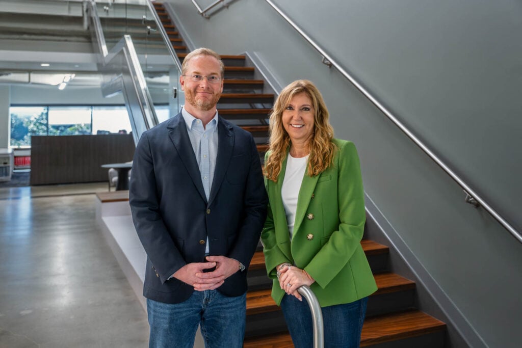 Two people stand indoors at the base of a staircase, both smiling at the camera. The setting appears to be a modern office building.