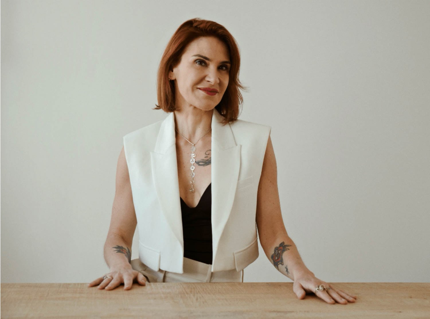 A woman with short brown hair, wearing a white sleeveless blazer over a black top, sits at a wooden table against a plain background. She has visible tattoos on her arms and chest.
