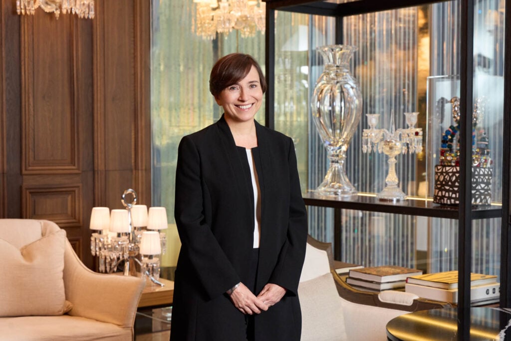 A woman in a black blazer stands smiling in a luxurious, well-lit room with crystal decor, books, and elegant furnishings.