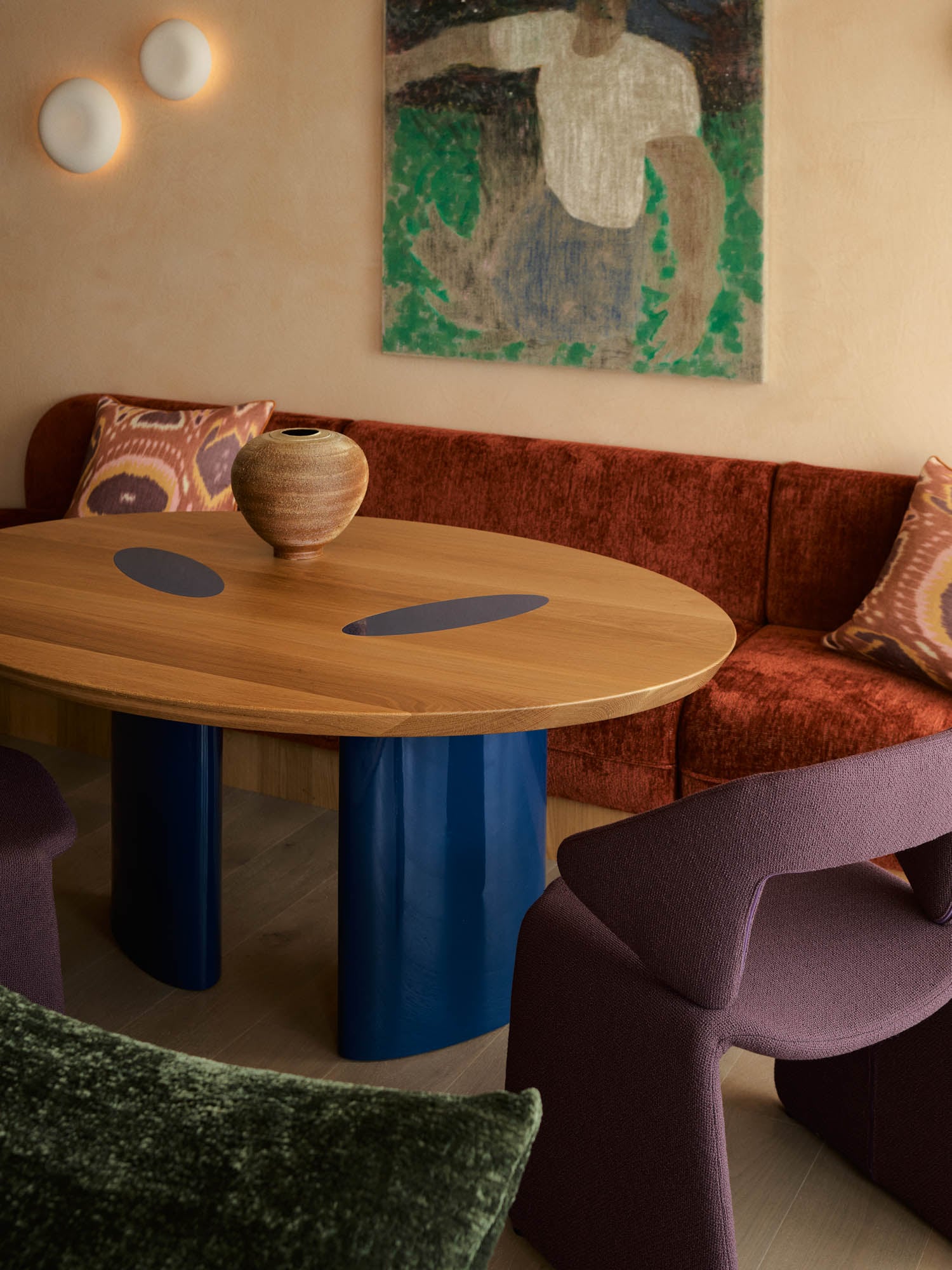 A modern dining area with a wooden table on blue legs, a red upholstered bench, purple chairs, decorative pillows, a ceramic vase, and abstract wall art.