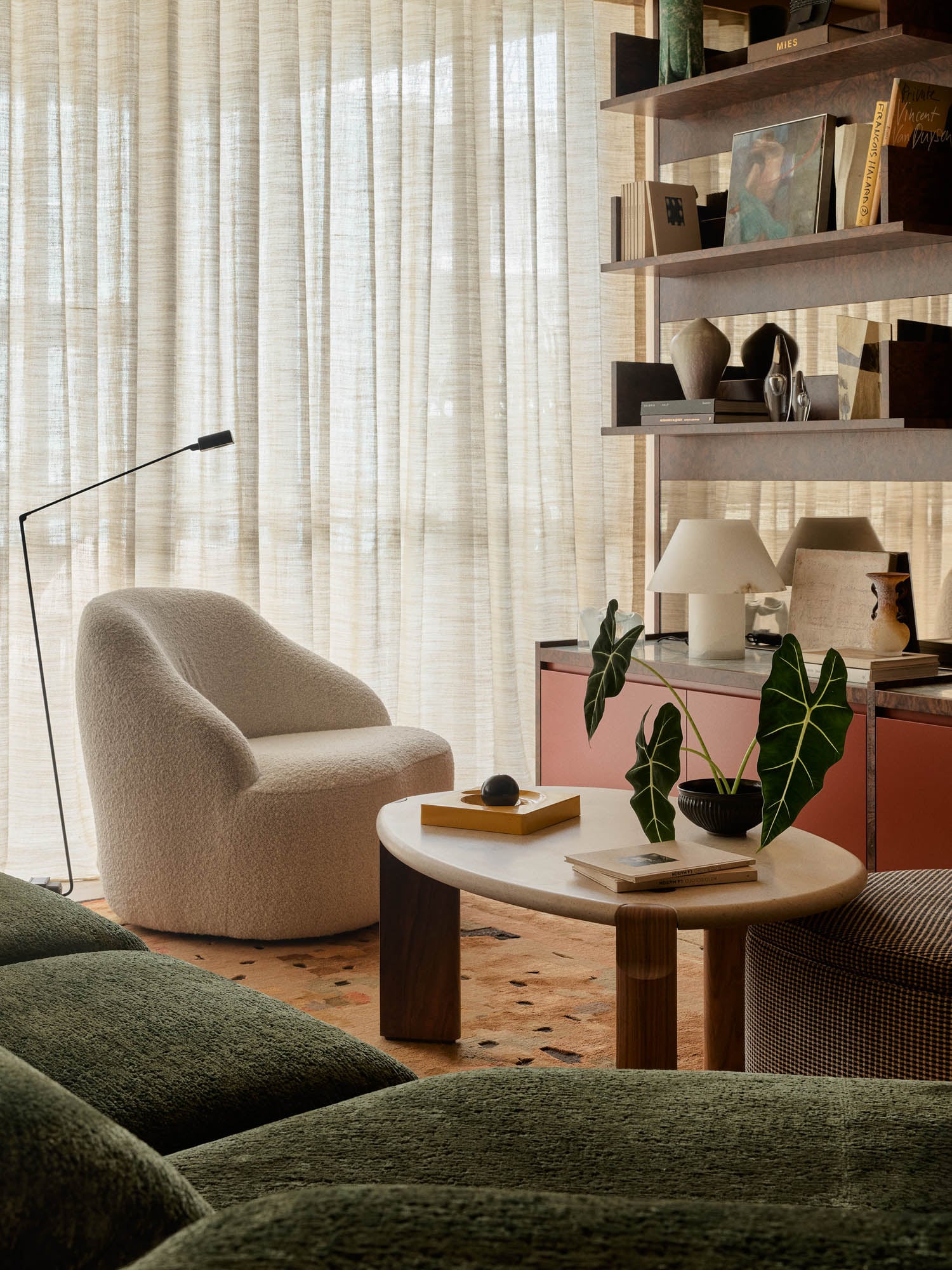 A cozy living room features a curved white armchair, a round table with books and a plant, green sofa, floor lamp, and a shelving unit with decor by a window with sheer curtains.