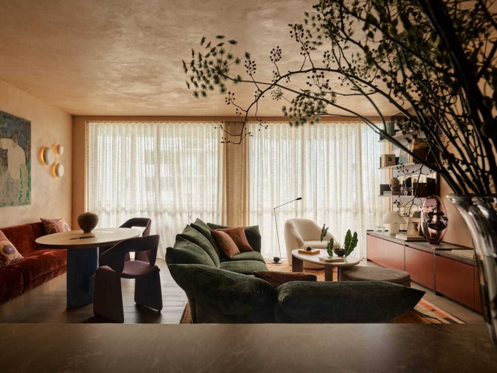 A stylish living room with large windows, sheer curtains, modern furniture, a green sofa, round dining table, red cabinet, and indoor plants.