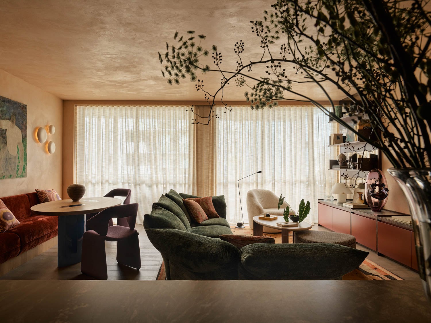 A stylish living room with large windows, sheer curtains, modern furniture, a green sofa, round dining table, red cabinet, and indoor plants.