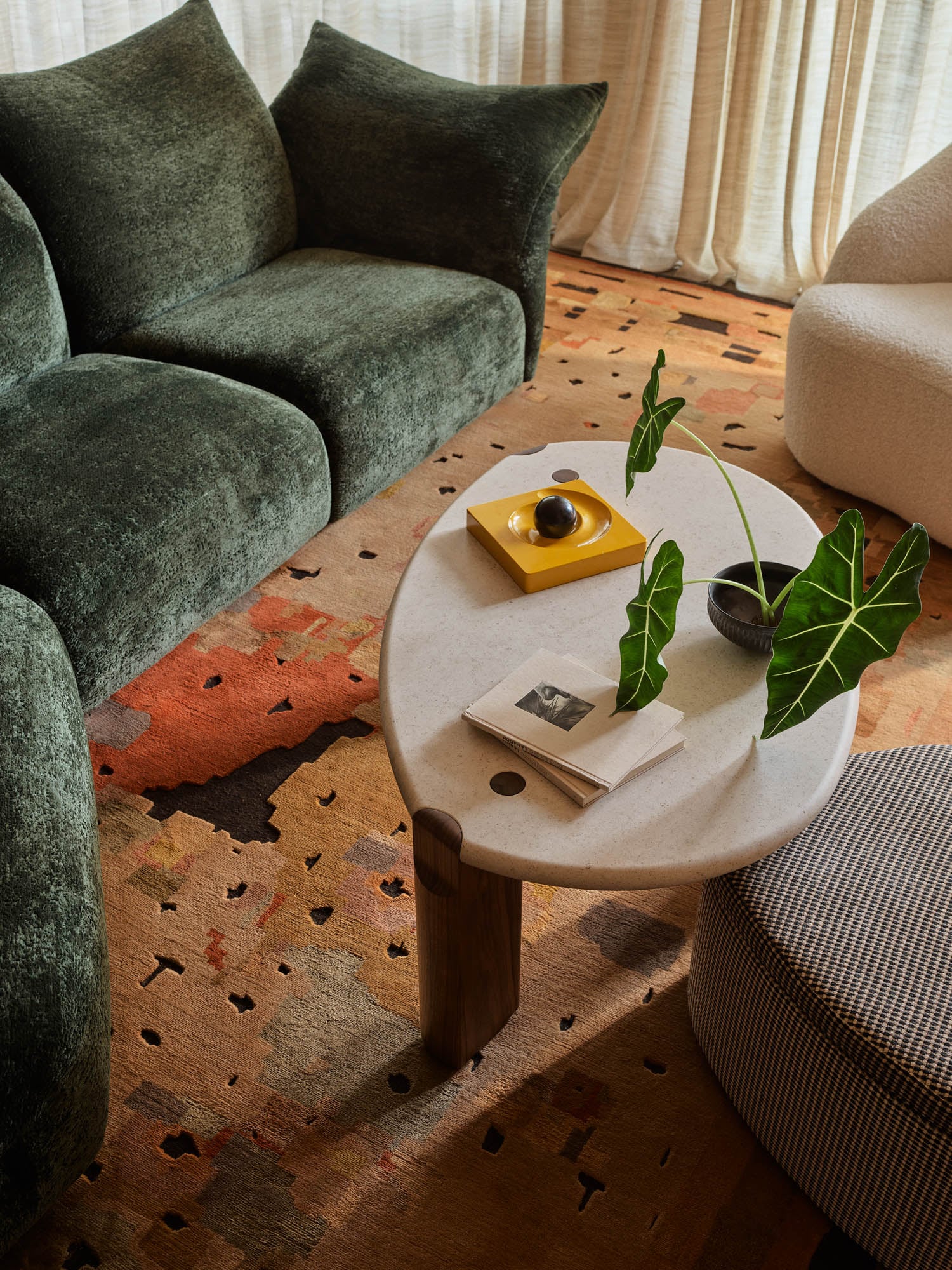A modern living room with a green velvet sofa, patterned rug, oval marble coffee table holding books, a yellow tray, a small bowl, and two leafy plants.