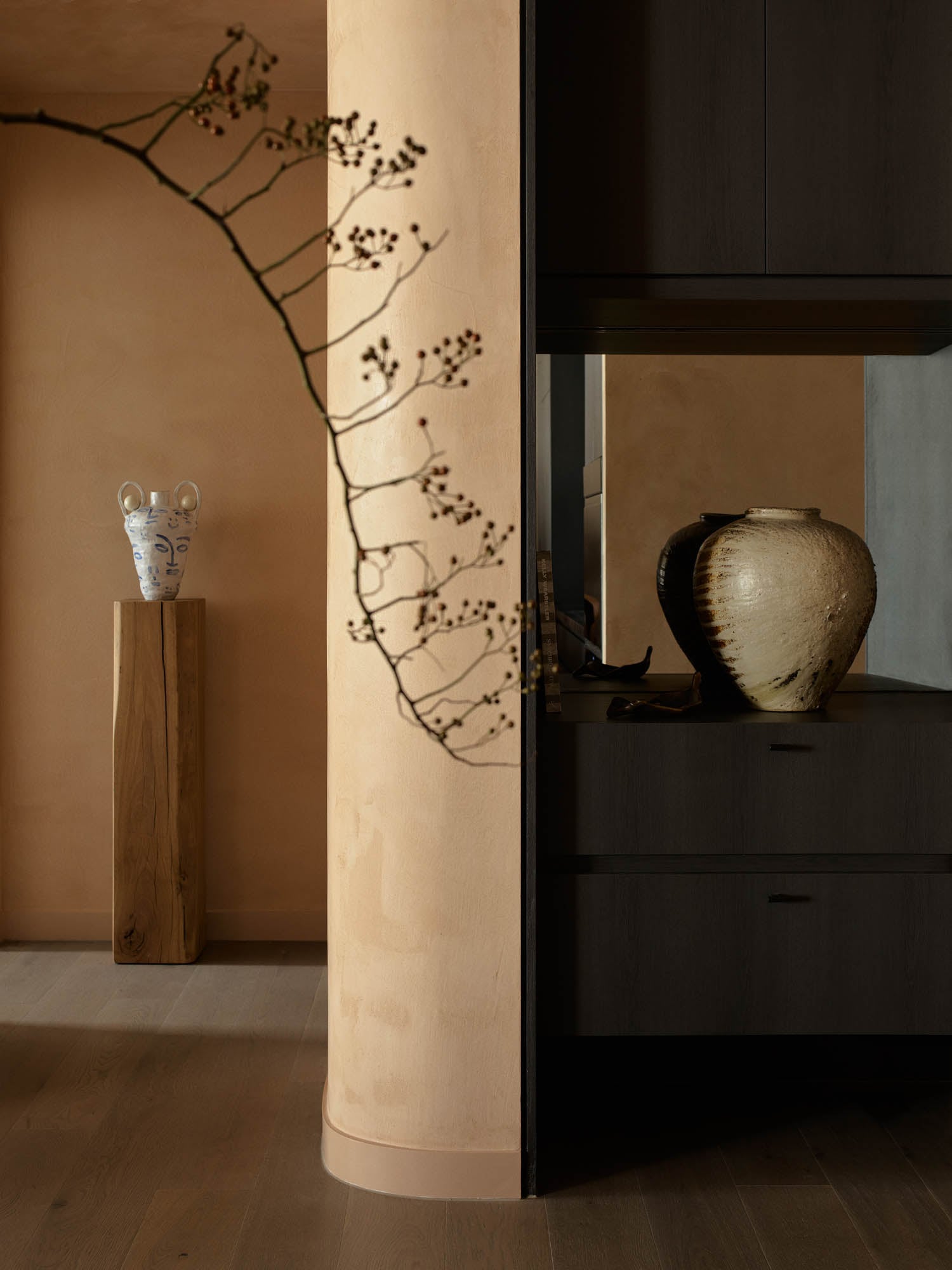 A minimalist interior features a beige curved wall, a wooden pedestal with a white vase, a dark cabinet, and a large pottery jar; a bare branch is visible in the foreground.