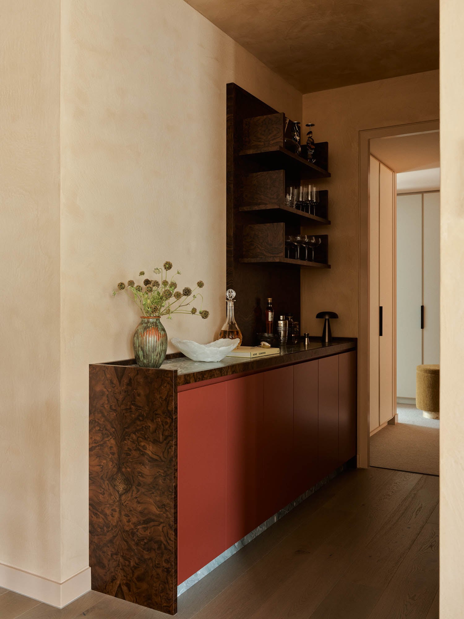 A modern hallway bar with red cabinets, dark wood shelving, glassware, bottles, a vase with flowers, a decanter, and neutral-toned walls and flooring.