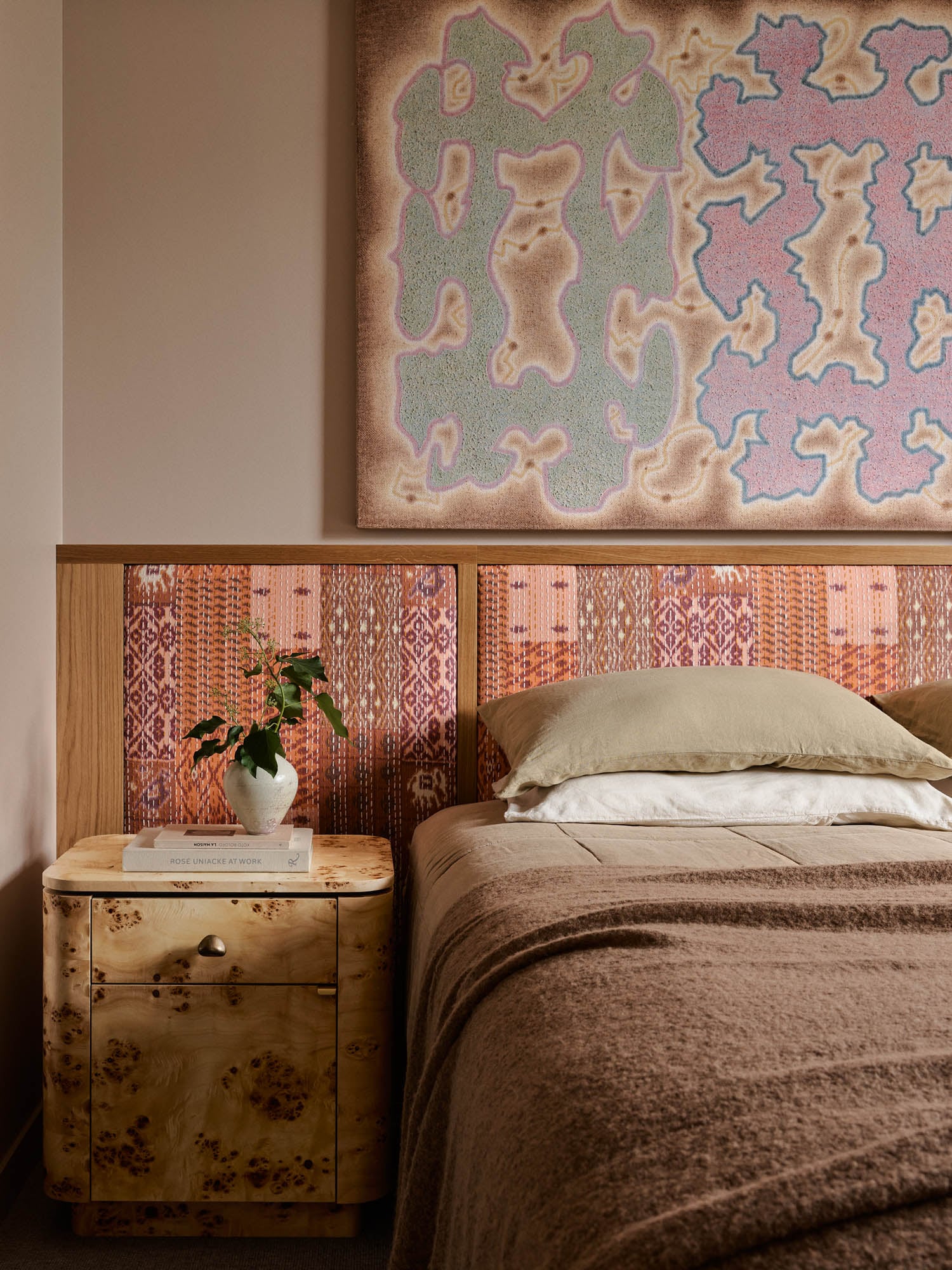 A neatly made bed with beige and brown bedding, a patterned headboard, a wooden nightstand with a book and vase, and abstract artwork on the wall above.
