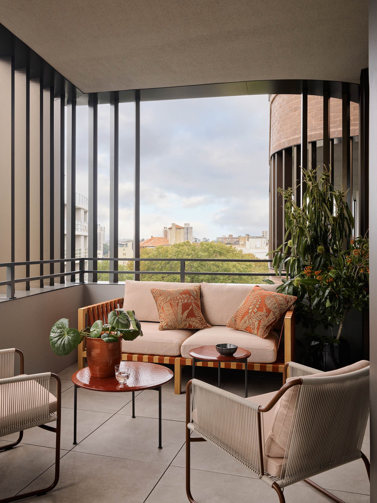 A modern balcony with a cushioned wooden sofa, two chairs, a round table with potted plants, and city buildings visible in the background.