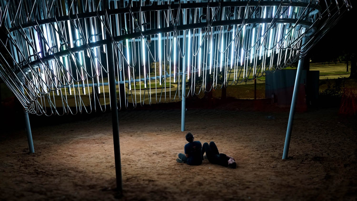 Two people lie on the ground at night under an illuminated art installation made of hanging light tubes in an outdoor area.