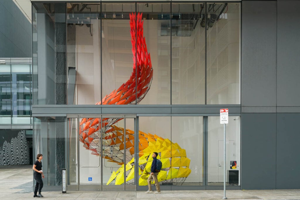 Two people walk past a glass-walled building displaying a large, colorful spiral sculpture with yellow, orange, and red angular segments inside.