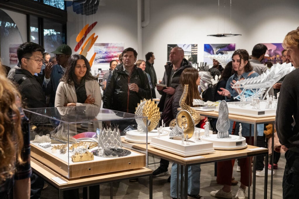 People view intricate architectural models displayed in glass cases at a busy indoor exhibition.